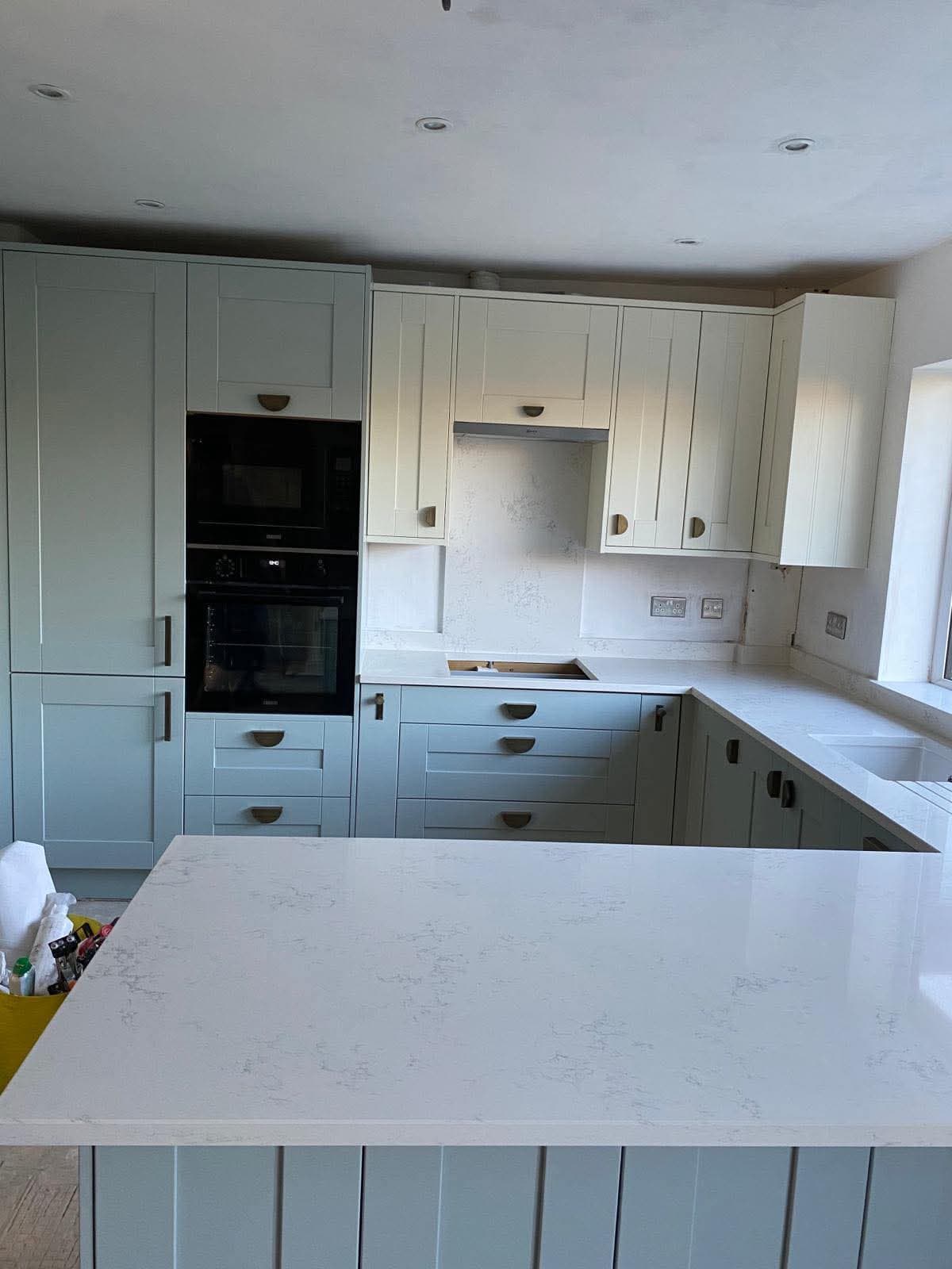 Installed fitted kitchen with pale blue-grey cabinetry, white quartz countertops, integrated black oven, and recessed ceiling lights.