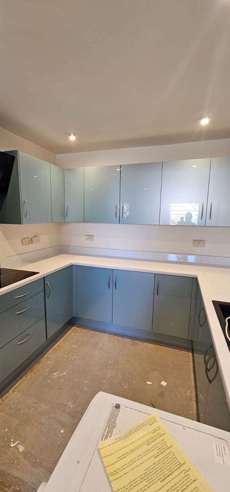 Installed fitted kitchen with soft blue-grey lower cabinets, white quartz countertops, frosted glass upper cabinets, downlights in white ceiling, and concrete subfloor during construction phase.