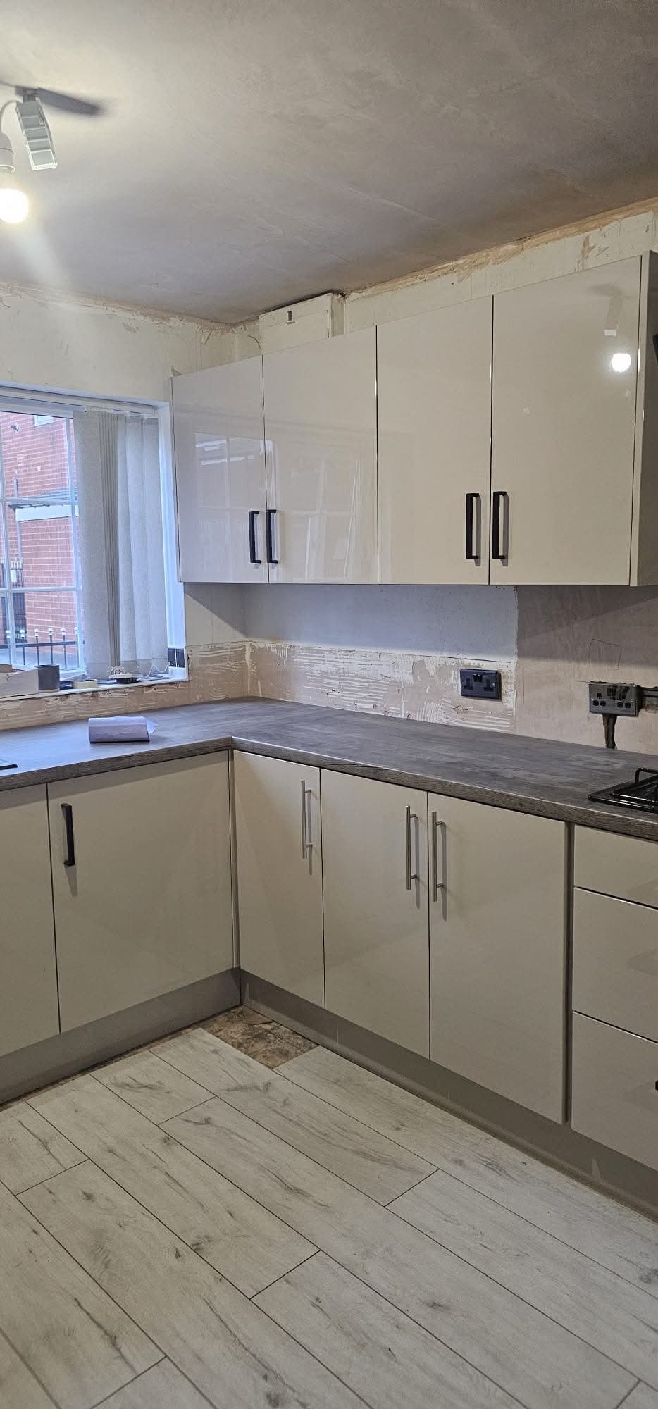 Cream and grey u-shaped kitchen with dark grey stone worktop, beige painted cabinets, grey tiled floor, and window view.