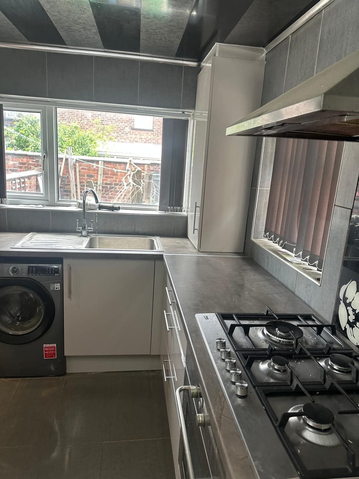 Cream and grey two-tone cabinetry with dark grey tiled backsplash, grey stone worktop, gas range cooker and integrated washing machine in a small galley kitchen with brick exterior visible through window.