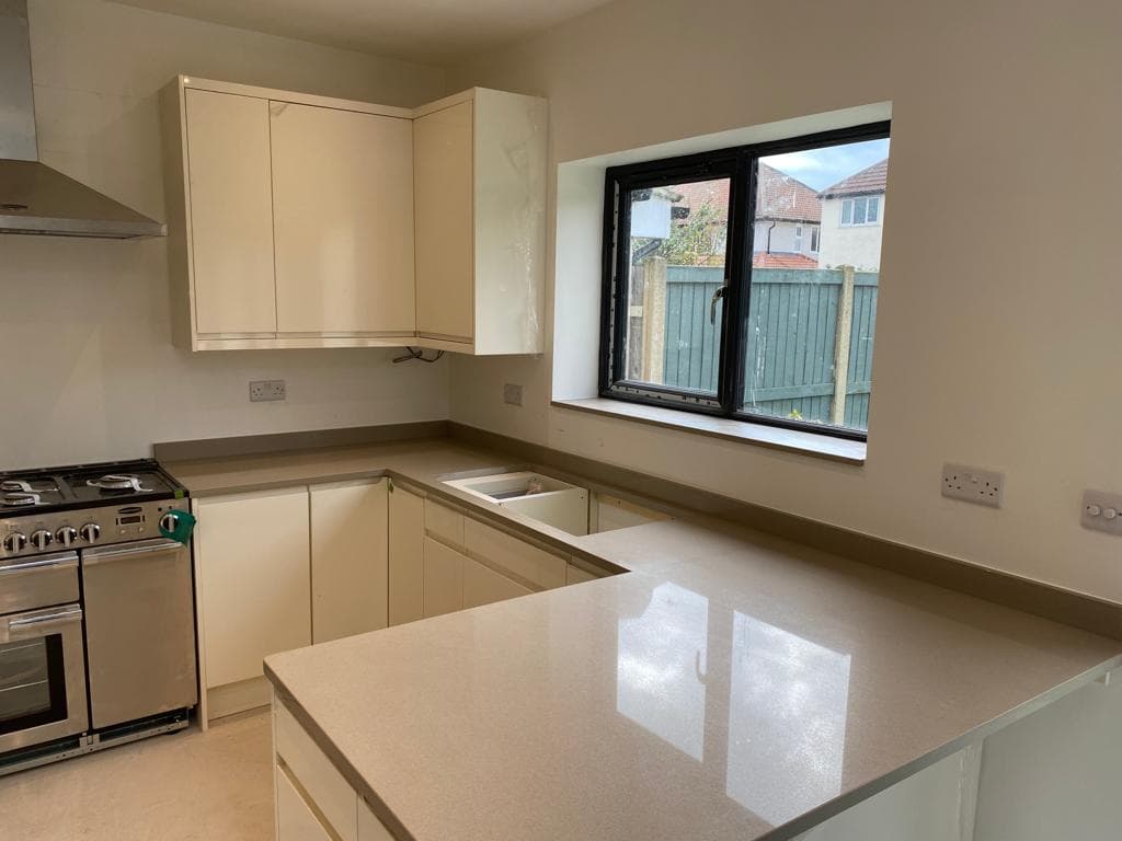 U-shaped kitchen with cream-coloured painted units, beige quartz worktops, stainless steel range cooker, and large black-framed window overlooking garden
