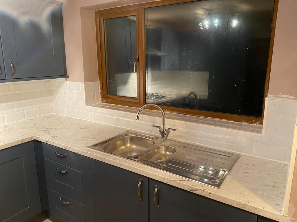 L-shaped kitchen with dark grey painted cabinets, light marble worktop, white subway tile splashback, and stainless steel double sink under a window with wooden frames.
