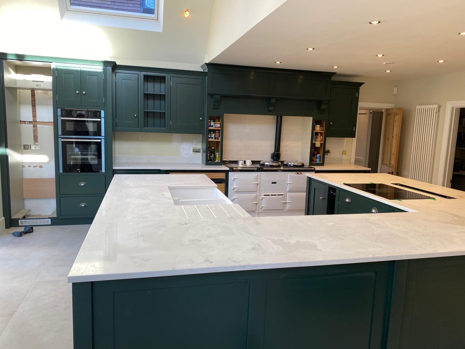 Large open-plan kitchen with dark green shaker-style cabinetry, light stone worktops, a central island with seating, and a cream Aga cooker, featuring pendant lights and integrated double oven.