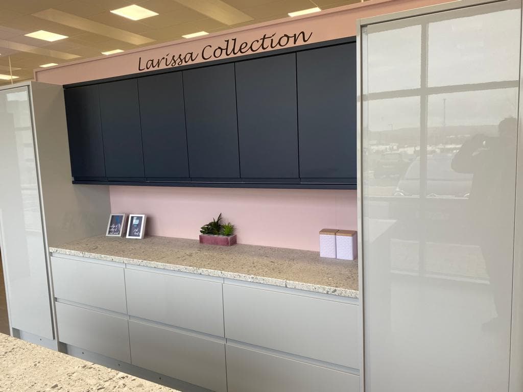Modern two-tone kitchen display with navy upper cabinets, white lower cabinets, and beige stone worktop against a pink feature wall.